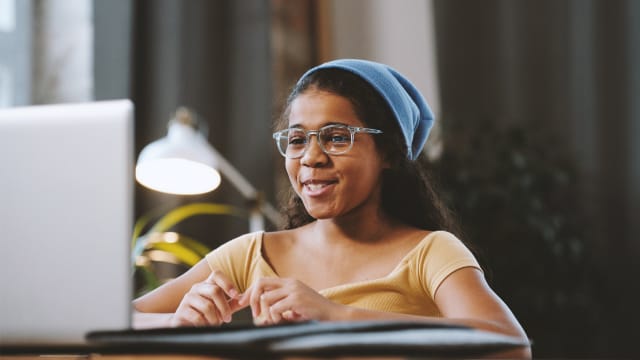 Ein junges Mädchen mit Brille, blauer Mütze und gelben T-Shirt sitzt im Arbeitszimmer vor einem Laptop am Tisch. Sie lächelt und redet dabei in die Kamera des Laptops.