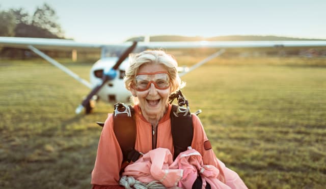 Eine ältere Dame kommt gerade vom Fallschirmspringen. Im Hintergrund ist das Flugzeug zu sehen. Es scheint die Sonne während sie auf einer Wiese steht.